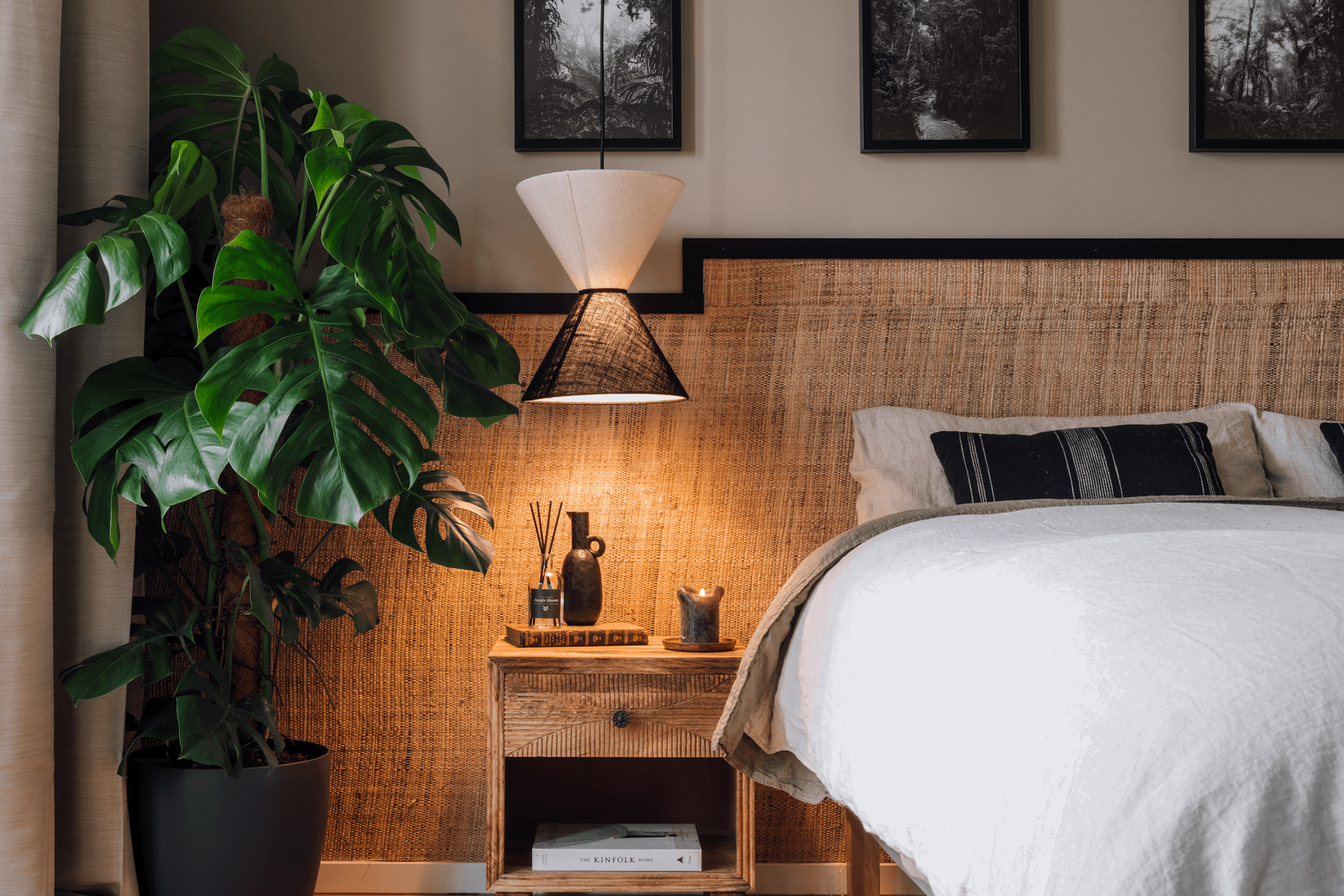 a bedroom with a bed, a nightstand and a potted plant. Monstera Deliciosa, linnen bed sheets, wooden nightstand with drawer, cozy bedroom with textured wallpaper, bamboo wallpaper, black photo frames, black and white pictures, lechuza plant pot. Aptó
