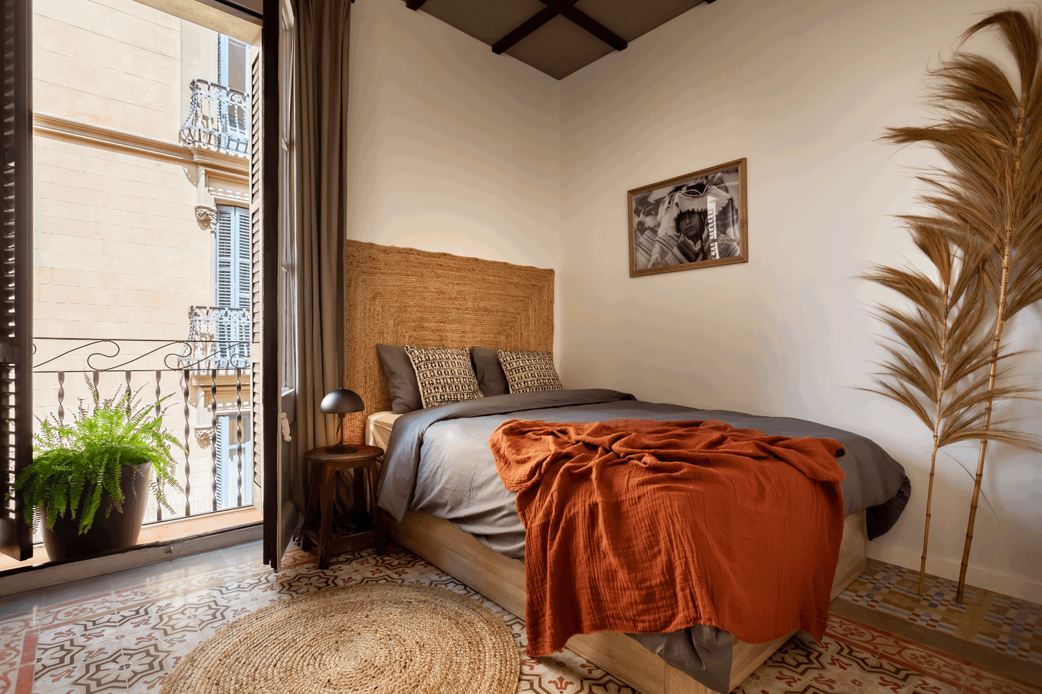 a bedroom with a bed, a rug and a window. Barcelona floor tiles, Barcelona interior design, painted ceiling, headboard design, wooden stool, black table lamp, linnen bed sheets, jute rug, best interior designer barcelona, Populair interior, Aptó 