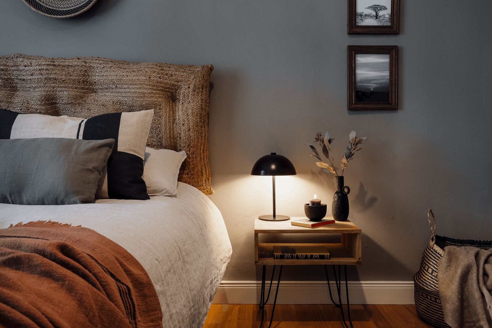a bedroom with a bed, a nightstand, and pictures on the wall. Small wall frames, Zara home interiors, cosy bed, pillows on bed, wooden night stand, big basked for bedroom, interior designer by Aptó interiors Berlin home design, Amsterdam homes