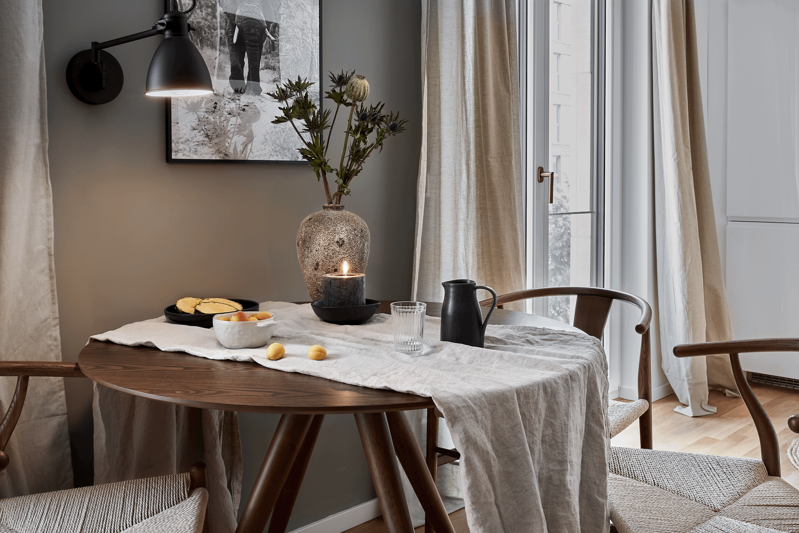 a small dining room with a table and chairs. Berlin interior design. Dark ooden round dining table, wishbone chairs, zara home interior, beige linnen curtains, grey wall paint, cozy home, dining area. Home staging. By Aptó interior, Noelle Baartmans 