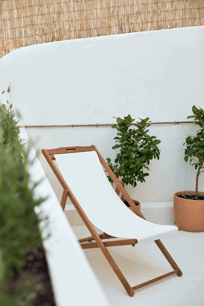 a lounge chair on a balcony with a potted plant next to it. Lemon tree and white canvas beach chair with wooden frame. white lime wash paint, interior design. Photoshoot staging, home staging, interior decorator. Designed by Aptó interiors.