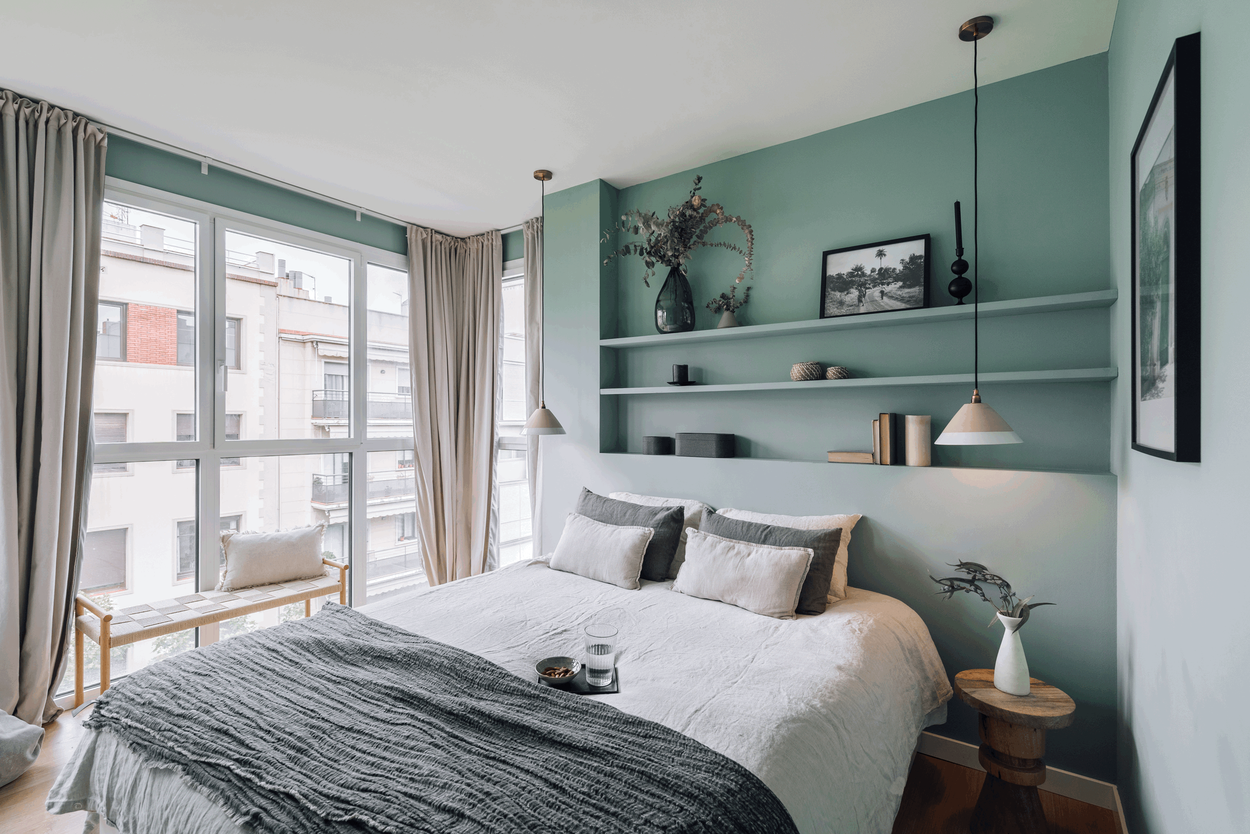 a bedroom with green walls and a bed. Minimalist cozy bedroom design. Cermic lamps, wooden stools, linnen sheets, zara home decoration. H&M home, linnen curtains, organic bedroom design. shelfs above headboard with decoration. By Aptó interiors