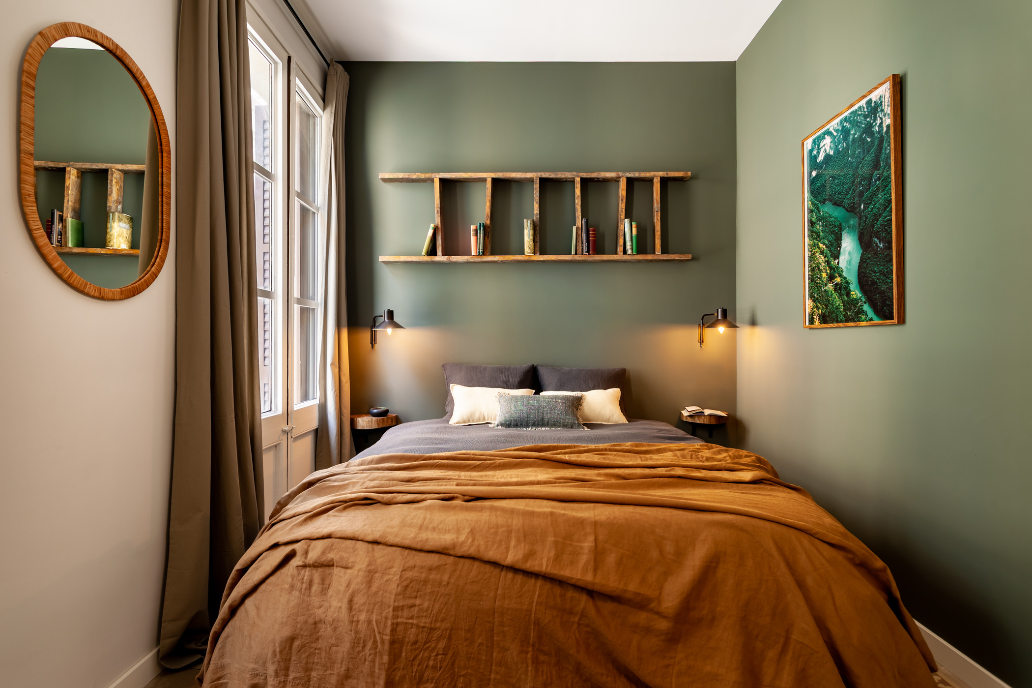 a bedroom with green walls and brown bedding. Green bedroom walls, cozy bedroom design, mirror in bedroom, oval shaped mirror, black wall lights, pillows on bed, blanket on bed. Barcelona Interior design, Amsterdam interior design, Aptó interiors