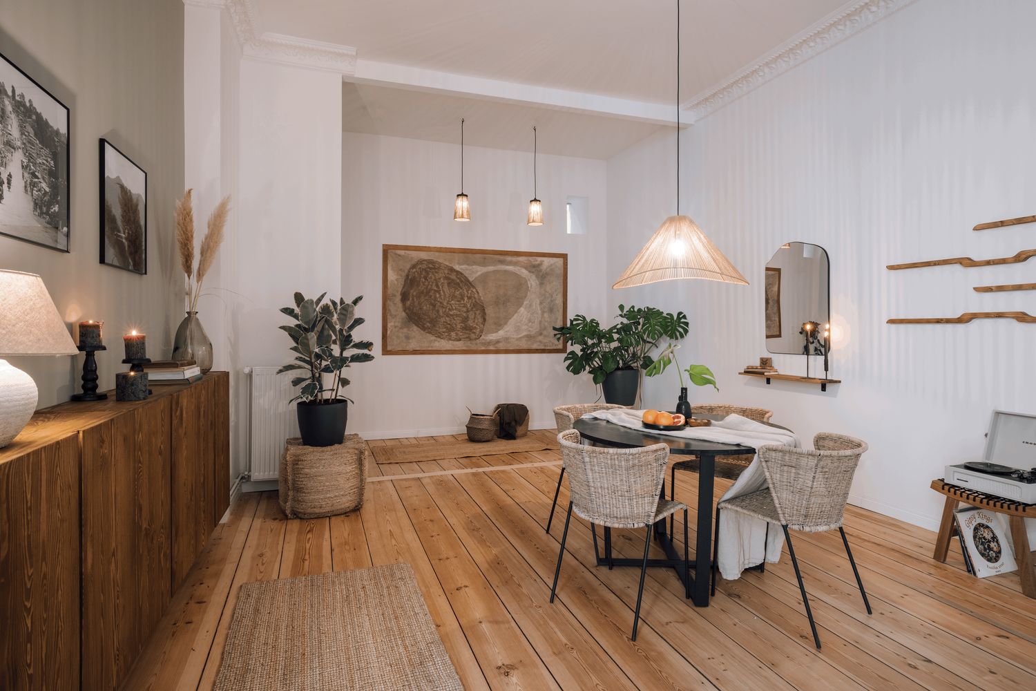 a dining room with wooden floors and white walls. Organic interior design, rattan chairs, organic shaped mirror, big abstract wall art with organic shapes in beige colours.  minimalist and cozy dining room setting with roudn table. by Aptó interiors